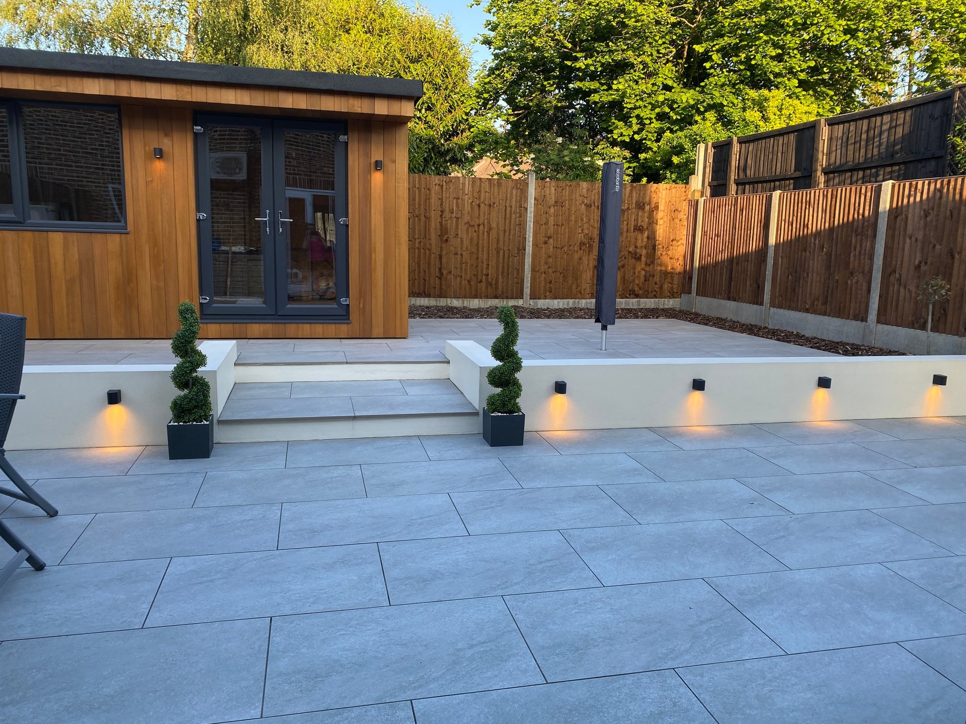 Modern garden patio with wooden garden studio, illuminated pathway lights, potted topiaries, and wooden fence at dusk