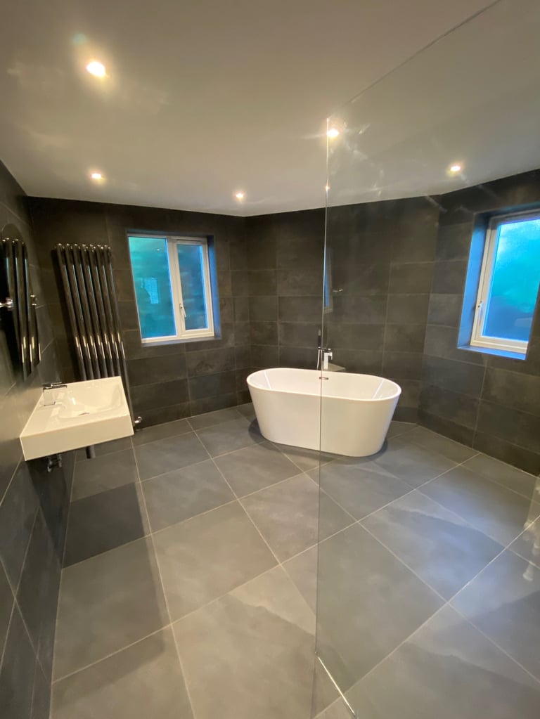 Modern bathroom with freestanding white bathtub, dark tile walls, recessed ceiling lights, and windows