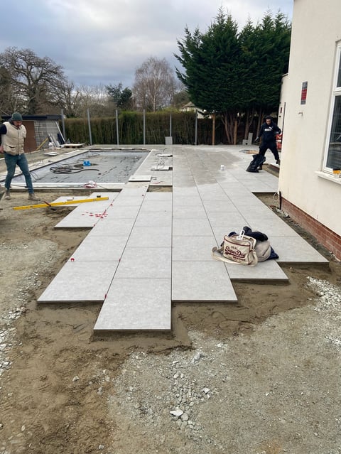 Construction workers installing large concrete paving stones on a residential patio, with trees and a white house visible in the background.