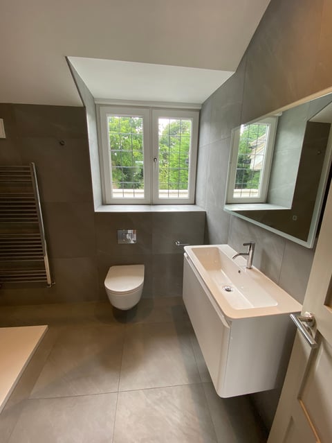 Modern bathroom with wall-mounted toilet, white sink vanity, large mirror, and windows overlooking green foliage