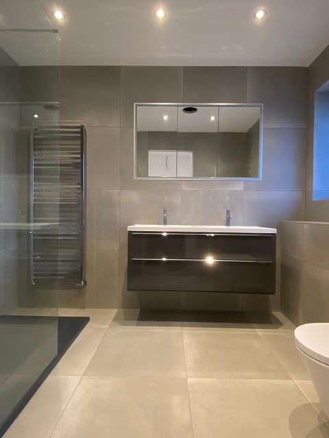 Modern bathroom with dark vanity, double sink, illuminated mirror, walk-in shower with glass enclosure, heated towel rack, and beige tile flooring