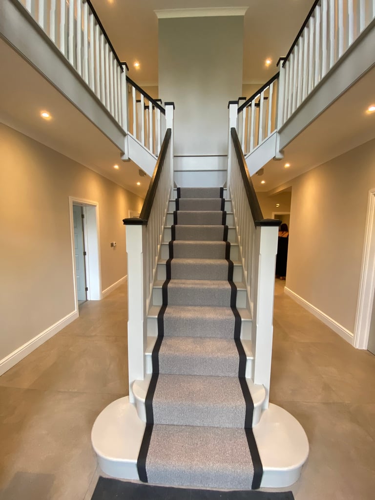 Modern staircase with gray carpet runner and black trim, flanked by white railings and warm-lit hallways on either side