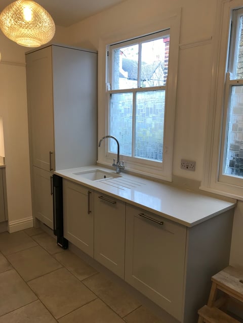 Modern white kitchen with sink and countertop beneath large window, brass pendant light overhead