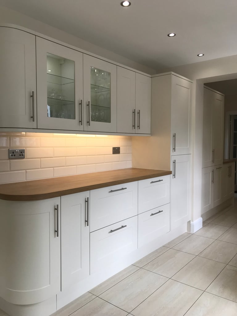 Modern white kitchen with wood countertop, glass-fronted cabinets, subway tile backsplash, and recessed lighting