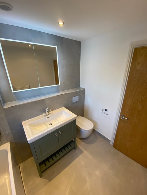 Modern bathroom with white sink, green vanity cabinet, illuminated mirror, toilet, and wooden door on light tile flooring