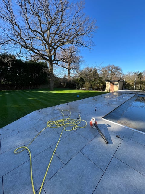 Worker installing blue patio tiles in a large backyard with bare trees and green lawn in the background on a sunny day