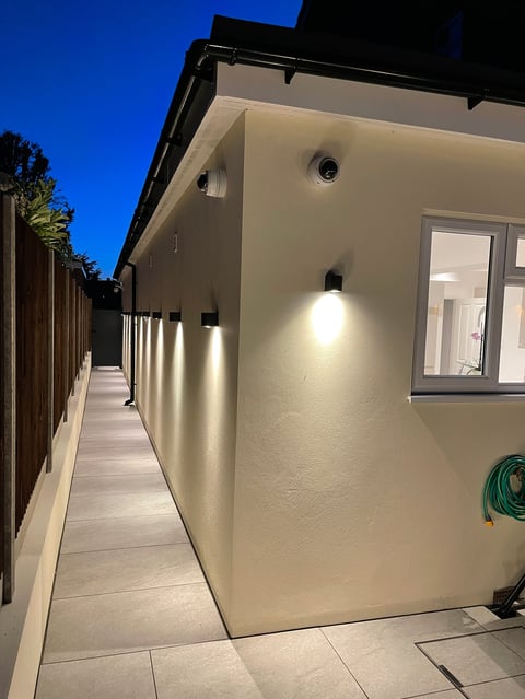 Modern cream-colored house exterior at dusk with wall-mounted lights, wooden fence, and paved pathway.