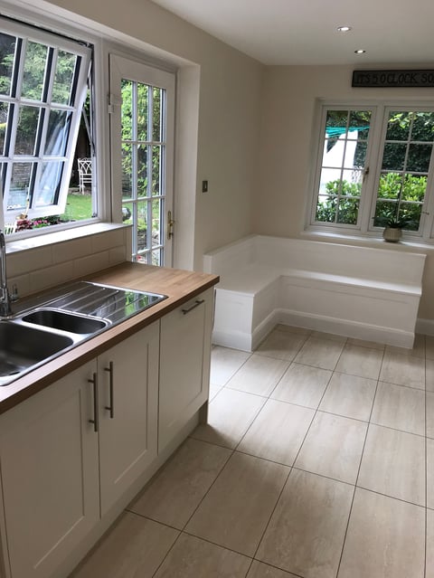 Modern kitchen with cream cabinets, wood countertop, sink near window, tiled flooring, and seating area with views of greenery outside