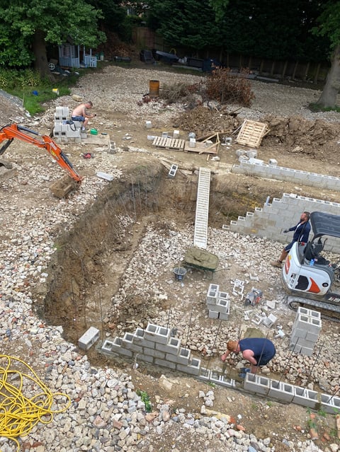 Aerial view of a construction site with excavated foundation, concrete blocks, gravel, and workers. Orange excavator and white mini excavator visible.