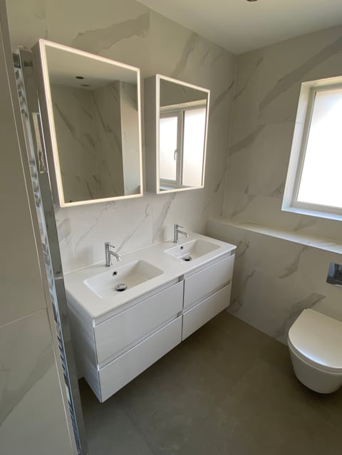 Modern bathroom with white dual-sink vanity, illuminated mirrors, marble tiles, window, and white toilet