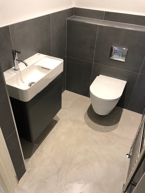 Modern bathroom with dark tile walls, white pedestal sink on left, white toilet on right, and beige concrete floor